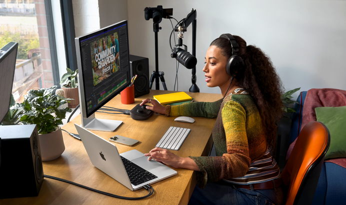 O femeie acasă lucrează la birou cu un MacBook Pro conectat la două monitoare externe, purtând căști și vorbind într-un microfon