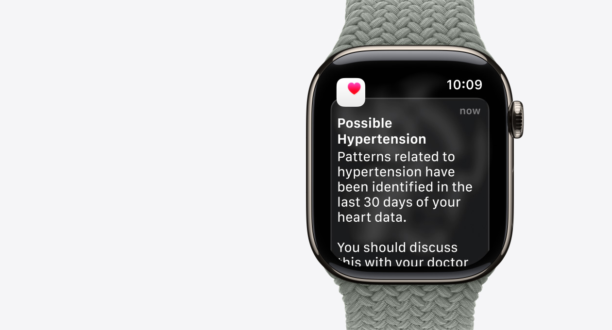 Apple Watch Series 11, титановий корпус натурального кольору, сповіщення про ймовірність гіпертензії, рожеве серце на тлі білого квадрата, з правого боку на годиннику розташована коронка Digital Crown, плетений монобраслет сіро-зеленого кольору